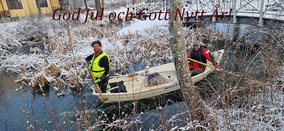 Foto på två personer i en roddbåt som tar sedimentprover i Jakob Mats damm, is, snö och en julhälsning.